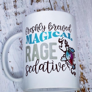 May include: White ceramic mug with the words "freshly brewed MAGICAL RAGE sedative" in a mix of fonts and colors. A colorful illustration of a potion bottle is on the right side of the mug. The mug has a white handle.