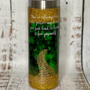May include: A gold glitter tumbler with a green and gold image of a yellow brick road. The tumbler has the quote "You've always had the power my dear you just had to learn it for yourself" printed on it.
