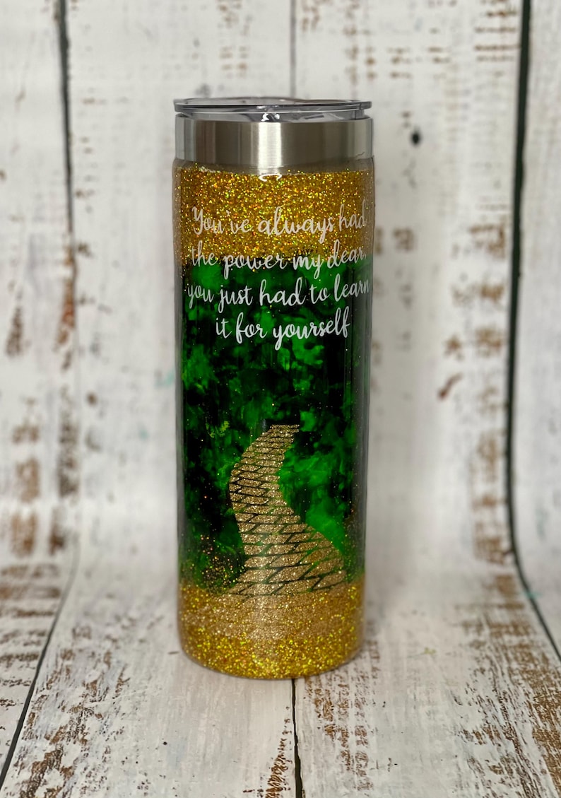 May include: A gold glitter tumbler with a green and gold image of a yellow brick road. The tumbler has the quote "You've always had the power my dear you just had to learn it for yourself" printed on it.