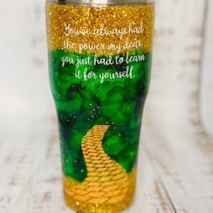 May include: A gold glitter tumbler with a green and gold design featuring a yellow brick road. The tumbler has the quote "You've always had the power my dear, you just had to learn it for yourself." printed on it.