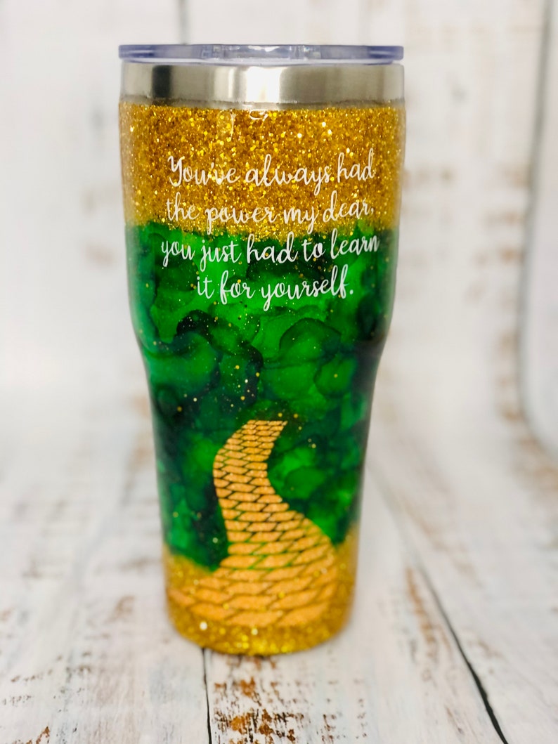 May include: A gold glitter tumbler with a green and gold design featuring a yellow brick road. The tumbler has the quote "You've always had the power my dear, you just had to learn it for yourself." printed on it.
