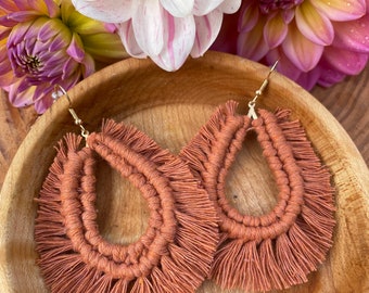 May include: A pair of terracotta-colored tassel earrings with a fringe design. The earrings are displayed on a wooden surface.