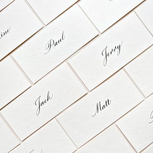 May include: A selection of white rectangular place cards, each bearing a name in elegant black calligraphy. The cards are arranged diagonally, creating a visually pleasing pattern. The names are written in a flowing script.