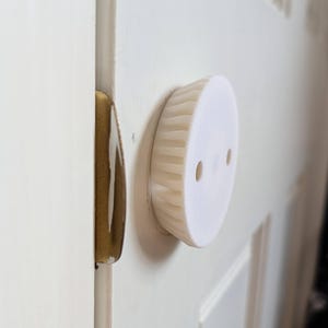 Op de afbeelding: Een witte, cirkelvormige deurbuffer met een geribbelde rand en twee kleine gaten, gemonteerd op een witte deur. De deur heeft een goudkleurige deurklink.