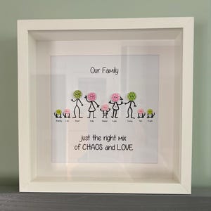 May include: A white framed family portrait. The artwork depicts stick figures with button heads, representing family members. The text reads "Our Family" and "just the right mix of CHAOS and LOVE."