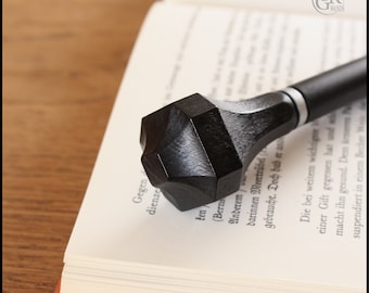 Puede incluir: Una varita de madera negra con un mango octogonal facetado. La varita está apoyada en un libro con texto en alemán.