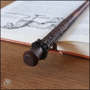 Könnte beinhalten: Ein dunkelbrauner Holzstab mit aufwendigen Schnitzereien und einer abgerundeten Spitze. Der Stab liegt auf einem Buch mit einer Schwarzweißillustration eines Pferdes.