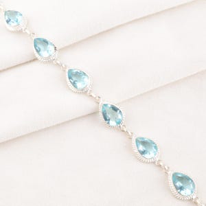 Op de afbeelding: Een zilveren armband met lichtblauwe, druppelvormige edelstenen. Elke edelsteen is gezet in een decoratieve zilveren bezel en verbonden met kleine zilveren connectoren. Op een witte stoffen achtergrond.