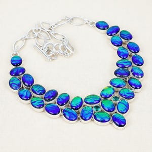 Könnte beinhalten: Eine silberne Halskette mit ovalen, irisierenden blau-grünen Steinen. Die Steine sind in silbernen Fassungen gefasst und in einem doppelreihigen Design angeordnet. Die Halskette hat einen Knebelverschluss und eine Kettenverlängerung.
