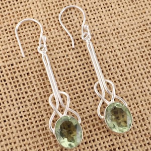 May include: A pair of silver-toned dangle earrings. Each earring features a long, slender design with a loop detail and a light green oval gemstone at the bottom. The earrings are displayed on a textured, tan surface.