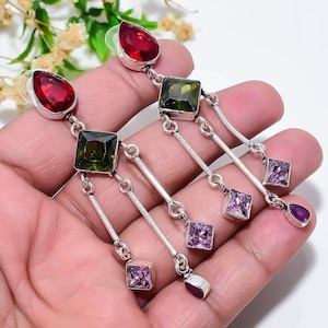 May include: A pair of silver dangle earrings featuring a teardrop-shaped red gemstone at the top, a square-cut olive green gemstone below, and three dangling silver chains ending in small square-cut light purple and teardrop-shaped red gemstones.