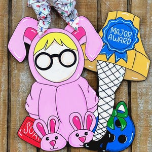 May include: A wooden cutout featuring a pink bunny suit with glasses, a leg lamp with "MAJOR AWARD" text, and a blue ornament. The bunny suit has a bow with Christmas lights. The cutout is on a wooden background.