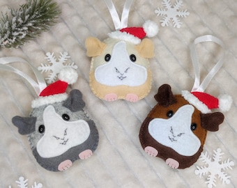 May include: Three felt guinea pig ornaments in shades of gray, brown, and beige, each wearing a red Santa hat with white trim. They have white faces and are hanging from white ribbons. White snowflake decorations and a small evergreen sprig are scattered around.