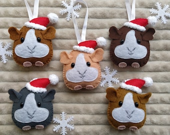 May include: Five felt guinea pig ornaments in various shades of brown and gray, each wearing a red Santa hat with a white pom-pom. They have white faces and are suspended by white ribbons. White snowflake decorations are scattered around.
