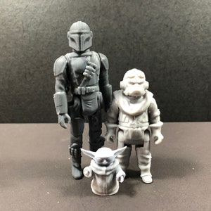 May include: Three gray action figures: a Mandalorian, a Grogu, and a character in a white spacesuit. The Mandalorian figure is the tallest, with a detailed helmet and armor. Grogu is in a robe. The figures stand on a dark surface.