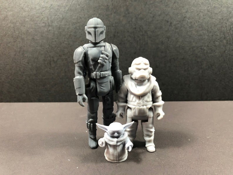 May include: Three gray action figures: a Mandalorian, a Grogu, and a character in a white spacesuit. The Mandalorian figure is the tallest, with a detailed helmet and armor. Grogu is in a robe. The figures stand on a dark surface.