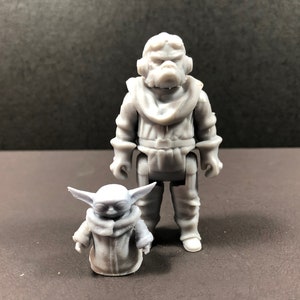 May include: Two gray action figures, one large and one small, stand side-by-side. The larger figure is humanoid with a detailed outfit. The smaller figure resembles a character with large ears and a hooded cloak. The figures are on a dark surface.