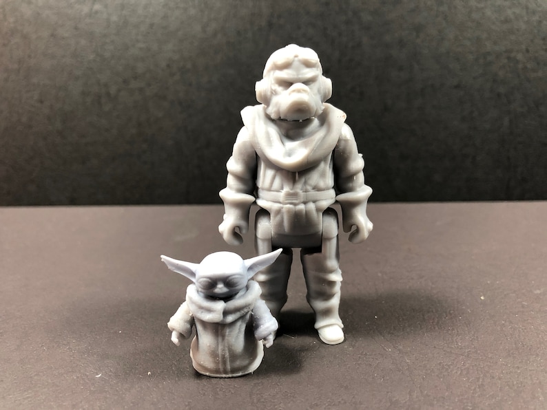 May include: Two gray action figures, one large and one small, stand side-by-side. The larger figure is humanoid with a detailed outfit. The smaller figure resembles a character with large ears and a hooded cloak. The figures are on a dark surface.
