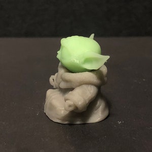 Pu&ograve; includere: Piccola figura verde chiaro di un personaggio con grandi orecchie e viso rugoso, sopra una figura grigia. La figura &egrave; una rappresentazione stilizzata di un personaggio popolare di un franchise di fantascienza.