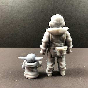 May include: A gray figurine set featuring two characters. One figure is a humanoid character in a hooded outfit, standing upright. The other is a small, green-skinned character with large ears, seated and facing forward. The figures are on a dark surface.