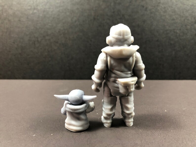 May include: A gray figurine set featuring two characters. One figure is a humanoid character in a hooded outfit, standing upright. The other is a small, green-skinned character with large ears, seated and facing forward. The figures are on a dark surface.