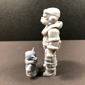 May include: A gray figurine of a character in a hooded jacket and a smaller creature. The larger figure is standing, and the smaller one is seated. The figures have a detailed, textured appearance.