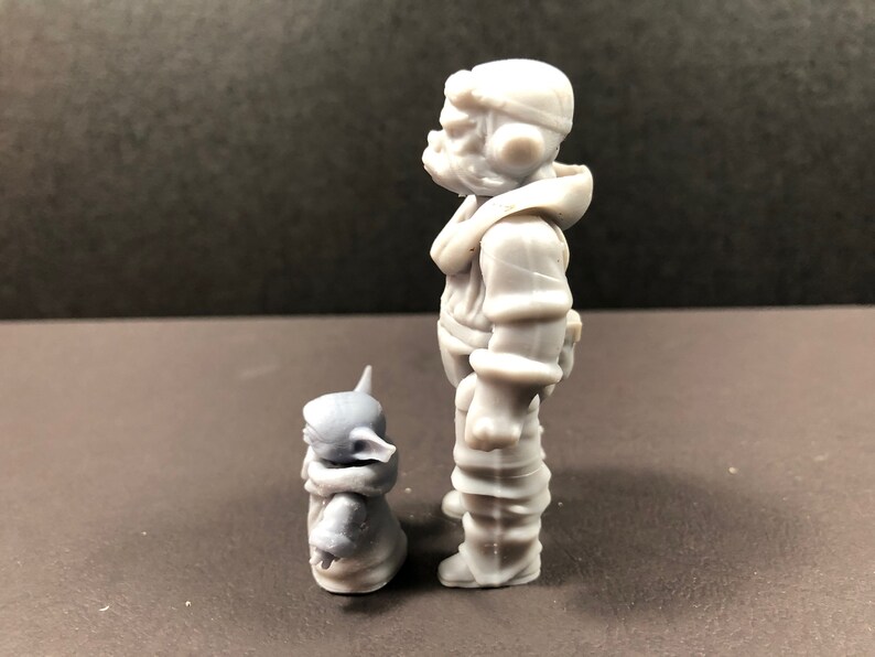 May include: A gray figurine of a character in a hooded jacket and a smaller creature. The larger figure is standing, and the smaller one is seated. The figures have a detailed, textured appearance.