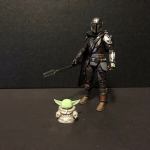 Pu&ograve; includere: Una dettagliata action figure di The Mandalorian, vestito con un'armatura scura e con un fucile blaster, &egrave; in piedi accanto a una piccola figura di Grogu. Le figure sono su una superficie scura, evocando una scena della serie Star Wars.