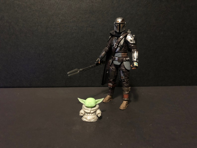 Pu&ograve; includere: Una dettagliata action figure di The Mandalorian, vestito con un'armatura scura e con un fucile blaster, &egrave; in piedi accanto a una piccola figura di Grogu. Le figure sono su una superficie scura, evocando una scena della serie Star Wars.