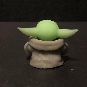 Pu&ograve; includere: Una piccola figura verde chiaro con grandi orecchie e una testa arrotondata, seduta in un indumento grigio con cappuccio. La figura &egrave; una rappresentazione stilizzata di un personaggio popolare di un franchise di fantascienza. La figura &egrave; su sfondo scuro.