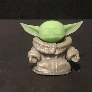Pu&ograve; includere: Piccola statuetta verde chiaro di un personaggio con grandi orecchie e una veste grigia. La statuetta &egrave; una rappresentazione stilizzata di un personaggio popolare di un franchise di fantascienza. La figura &egrave; su sfondo scuro.