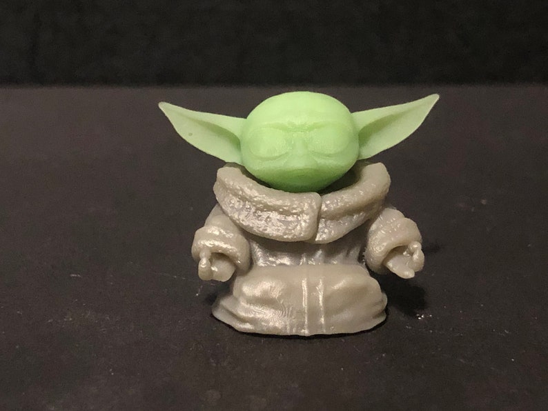 Pu&ograve; includere: Piccola statuetta verde chiaro di un personaggio con grandi orecchie e una veste grigia. La statuetta &egrave; una rappresentazione stilizzata di un personaggio popolare di un franchise di fantascienza. La figura &egrave; su sfondo scuro.
