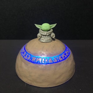 Pu&ograve; includere: Un oggetto decorativo a forma di cupola, marrone chiaro, con una fascia blu luminosa al centro. Una piccola figura verde con grandi orecchie &egrave; in cima. Probabilmente una luce notturna a tema o un pezzo decorativo.