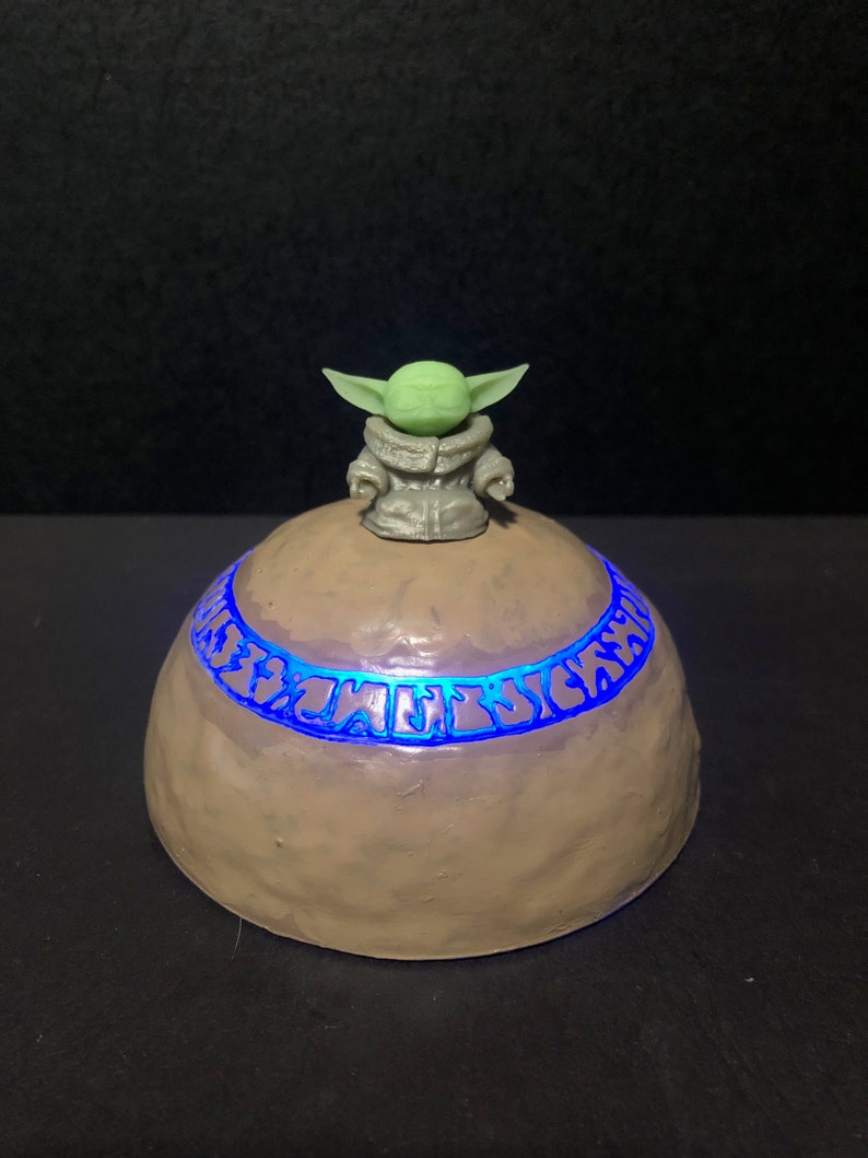 Pu&ograve; includere: Un oggetto decorativo a forma di cupola, marrone chiaro, con una fascia blu luminosa al centro. Una piccola figura verde con grandi orecchie &egrave; in cima. Probabilmente una luce notturna a tema o un pezzo decorativo.