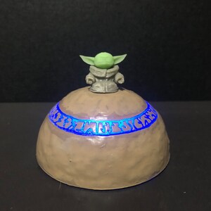 Pu&ograve; includere: Un oggetto decorativo artigianale a forma di cupola, marrone chiaro, con una fascia di testo blu brillante. Una piccola figura di personaggio dalla pelle verde &egrave; posta sopra la cupola. Probabilmente un oggetto da collezione o decorativo.