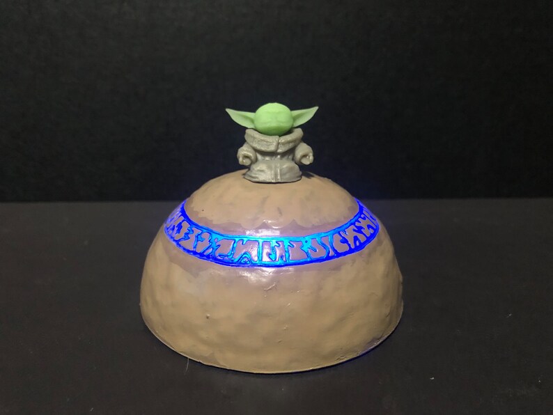 Pu&ograve; includere: Un oggetto decorativo artigianale a forma di cupola, marrone chiaro, con una fascia di testo blu brillante. Una piccola figura di personaggio dalla pelle verde &egrave; posta sopra la cupola. Probabilmente un oggetto da collezione o decorativo.
