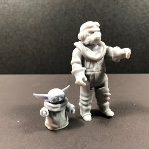 May include: Two gray figurines of characters from a science fiction franchise. One is a large humanoid figure in a spacesuit, and the other is a small, green-skinned alien with large ears. The figures are standing on a dark surface.