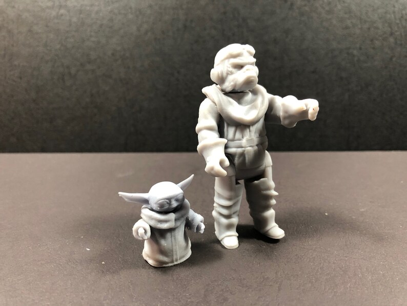 May include: Two gray figurines of characters from a science fiction franchise. One is a large humanoid figure in a spacesuit, and the other is a small, green-skinned alien with large ears. The figures are standing on a dark surface.