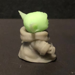 Pu&ograve; includere: Piccola statuetta verde chiaro di un personaggio con grandi orecchie e viso rugoso, che indossa una veste beige. La statuetta &egrave; in posizione seduta, con la testa e le orecchie ben in mostra. &Egrave; un oggetto da collezione.