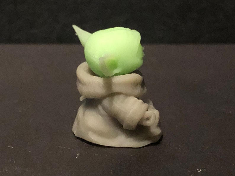 Pu&ograve; includere: Piccola statuetta verde chiaro di un personaggio con grandi orecchie e viso rugoso, che indossa una veste beige. La statuetta &egrave; in posizione seduta, con la testa e le orecchie ben in mostra. &Egrave; un oggetto da collezione.