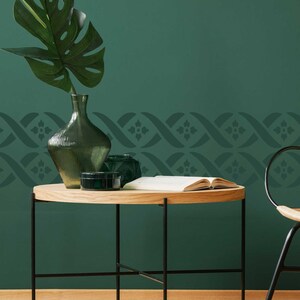 May include: A dark green wall with a decorative stencil border. A wooden table with black metal legs sits in front of the wall. A green glass vase with a leafy plant sits on the table.