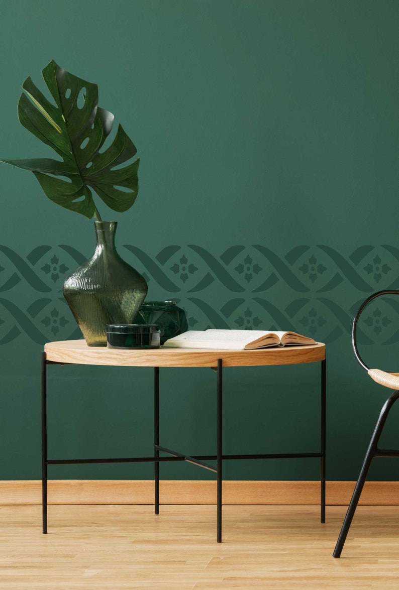 May include: A dark green wall with a decorative stencil border. A wooden table with black metal legs sits in front of the wall. A green glass vase with a leafy plant sits on the table.