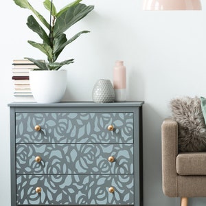 May include: A grey chest of drawers with three drawers features a floral pattern and gold knobs. The dresser is decorated with a potted plant, a vase, and a decorative object.