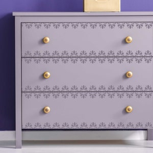 May include: A light purple chest of drawers with three drawers and gold knobs. Each drawer has a decorative stencil design around the edge.