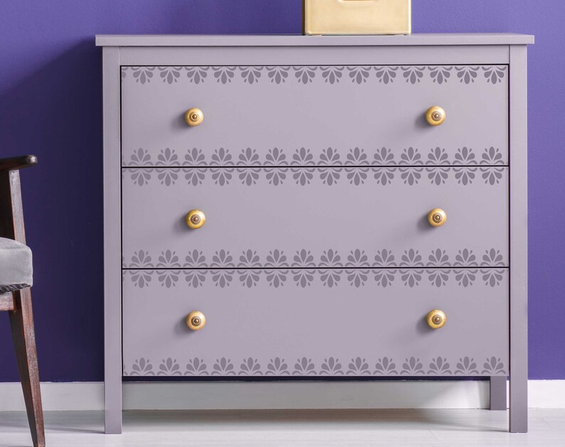 May include: A light purple chest of drawers with three drawers and gold knobs. Each drawer has a decorative stencil design around the edge.