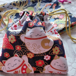 Może przedstawiać: Woreczek ze sznurkiem z japońskim wzorem kota Maneki Neko. Woreczek ma granatowe tło z czerwonymi i złotymi kwiatowymi akcentami oraz białymi ilustracjami szczęśliwych kotów. Sznurek jest złoty.