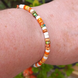 Peut inclure: Un bracelet coloré composé de petites perles carrées dans des tons de blanc, orange et vert. Le bracelet est porté au poignet.