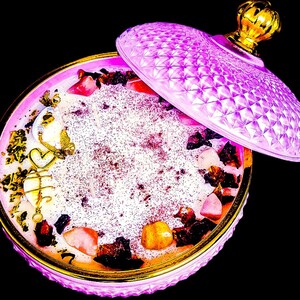 May include: A round, pink candle with a decorative lid, featuring a gold rim and accents. The candle is adorned with crystals, dried botanicals, and gold charms. The lid is open, revealing the candle's surface.