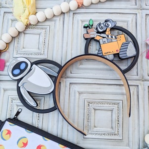 May include: A pair of black mouse ears with a white and grey robot character on one ear and a yellow robot character with the text "WALL-E" on the other ear.