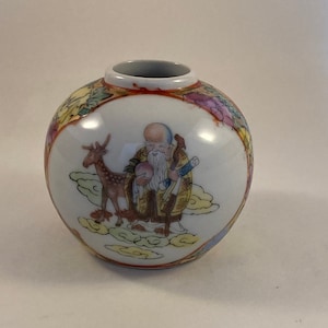 May include: A white porcelain vase with a round body and a red rim. The vase is decorated with a colorful image of a man with a long white beard and a deer, surrounded by clouds. The man is holding a peach and a scroll.